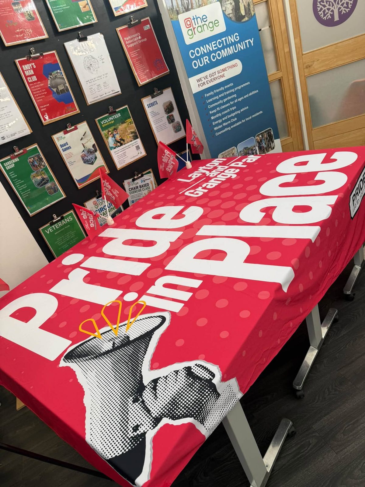 Pride in Place Layton and Grange Park branded table and flags in front of the @theGrange noticeboard